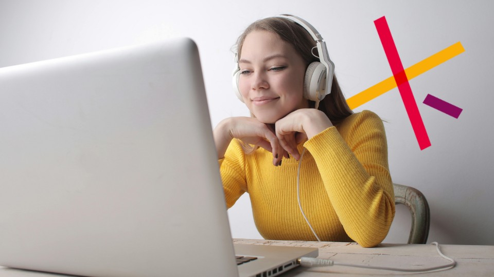 Keltapaitainen nainen katsoo hymyillen tietokonetta kuulokkeet päässään